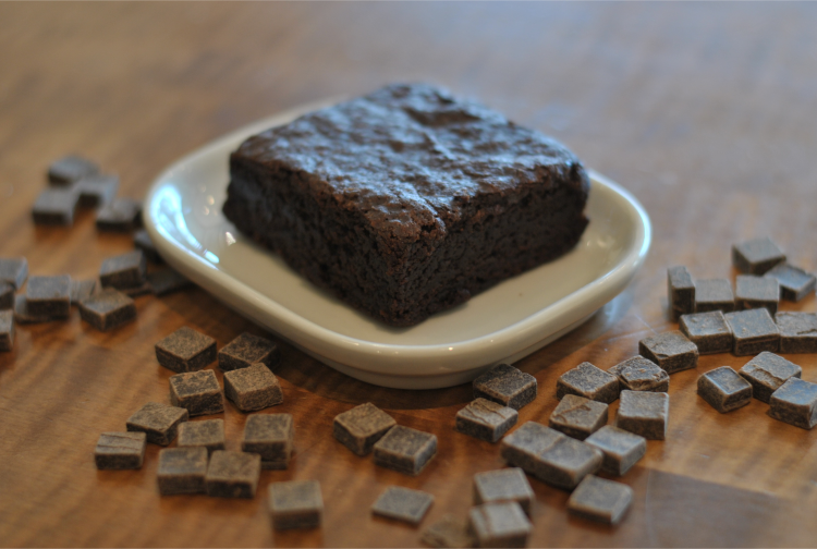 A dark chocolate brownie rests upon a small white plate, surrounded by chips of chocolate.