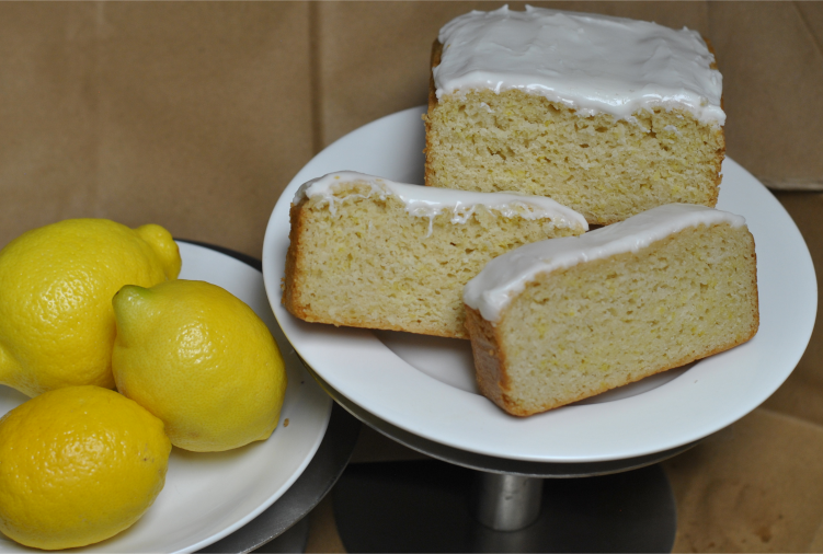 Bright iced lemon loaf slices rest upon a plate. Fresh lemons sit beside the plate. 