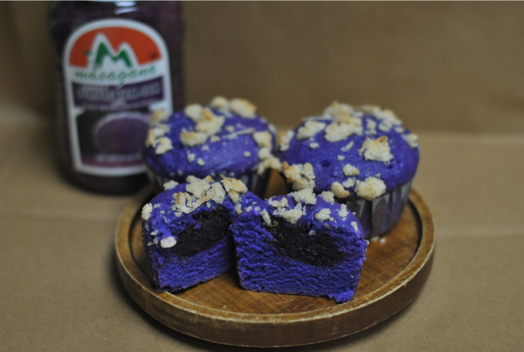 Bright purple muffins adorn a wooden plate. The sliced muffin in front shows a jammy ube center. 